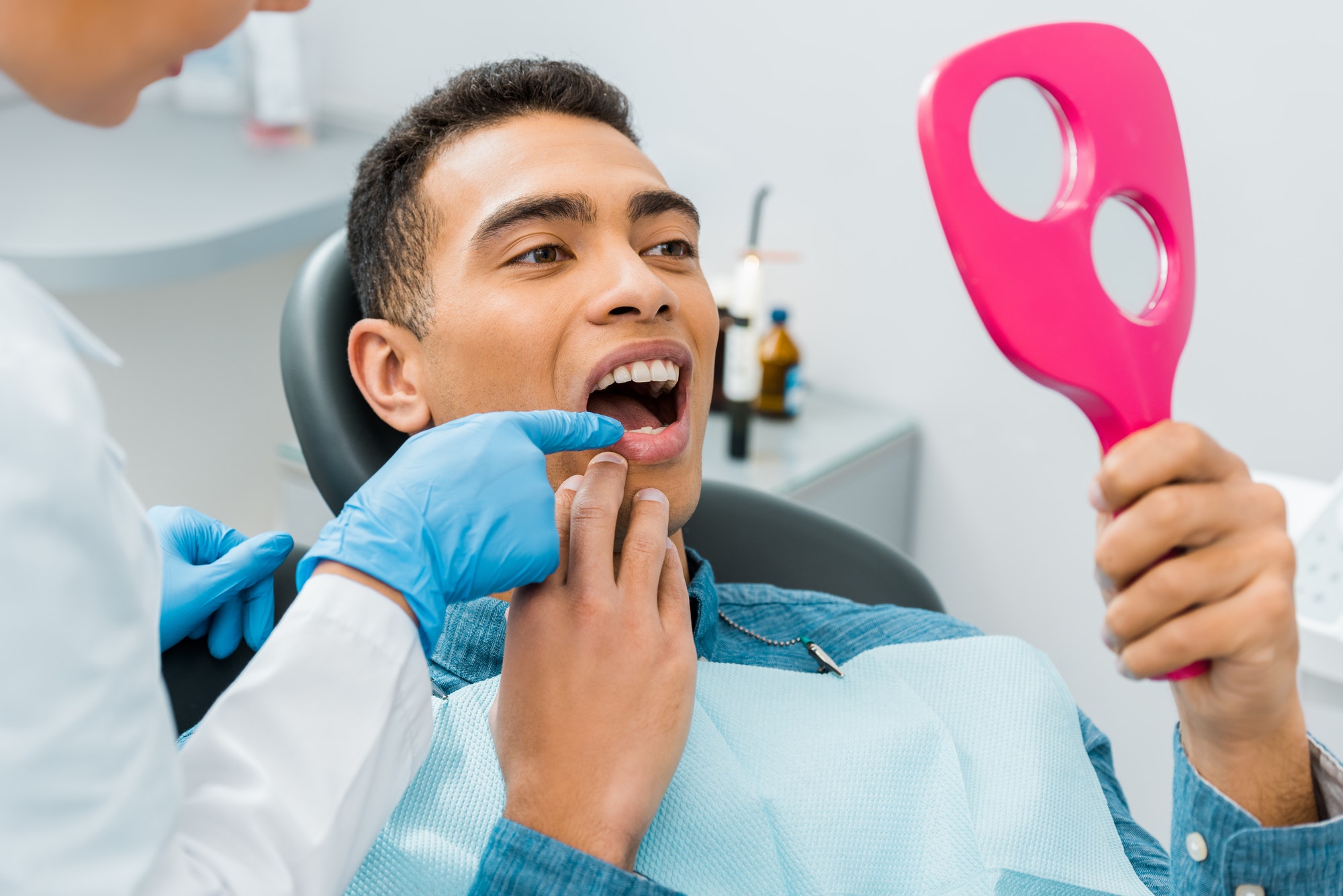 female dentist touching tooth of handsome african american man looking at mirror